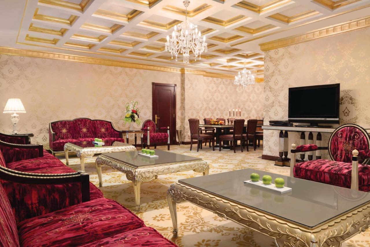 Grand Regency Doha, Trademark Collection By Wyndham Екстериор снимка The photo depicts an elegantly designed living room. The room features luxurious red sofas with ornate designs and metallic gold accents, arranged around glass-topped coffee tables that have intricate bases. A large, modern flat-screen television is 