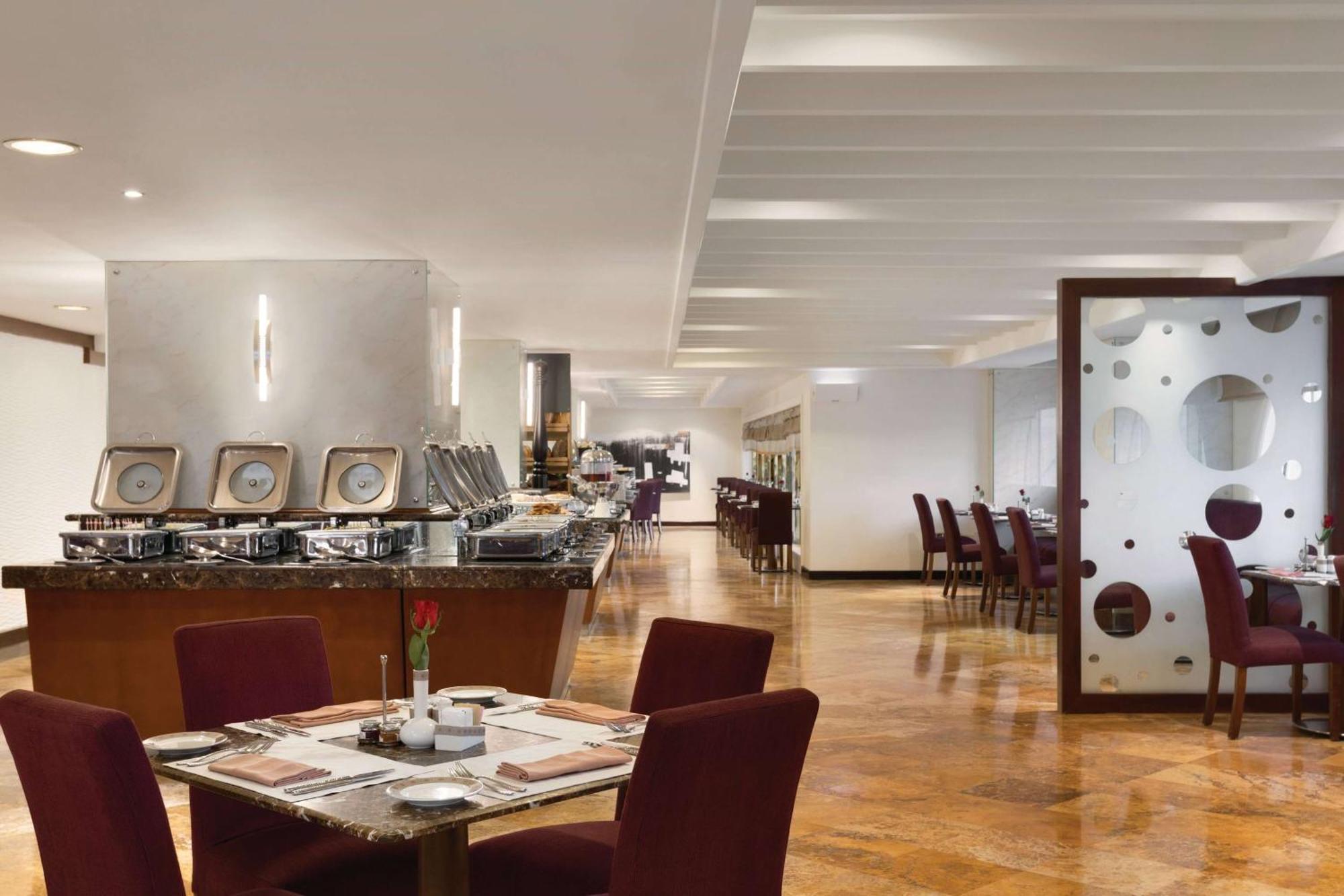 Grand Regency Doha, Trademark Collection By Wyndham Екстериор снимка The photo shows a modern restaurant interior. There are several tables with maroon chairs arranged throughout the space. One table is set with a small vase containing a flower and dining utensils. Along one side, there is a buffet area with a sleek c