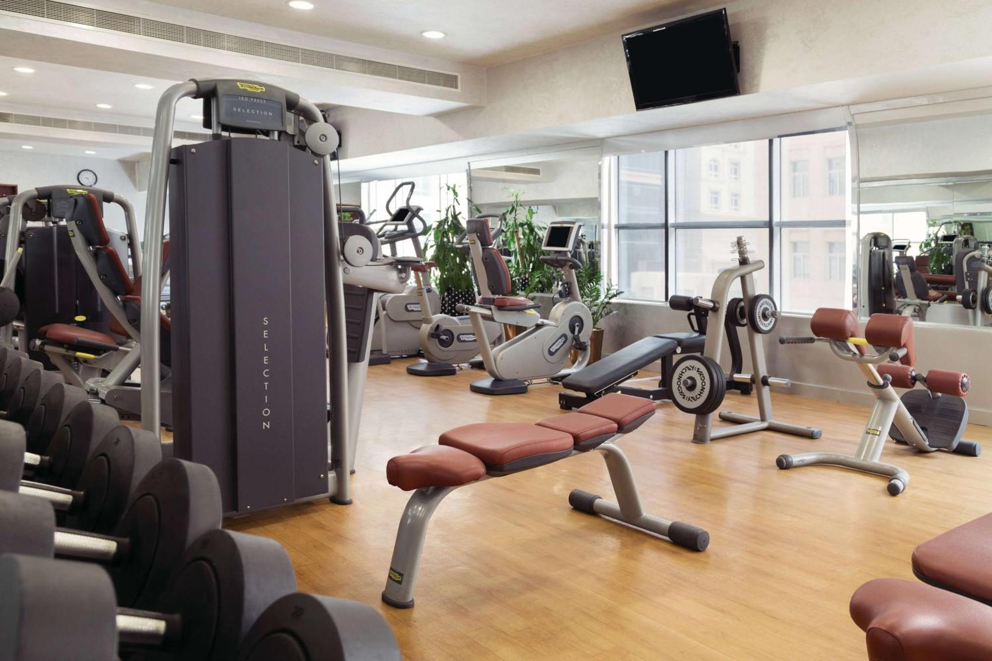 Grand Regency Doha, Trademark Collection By Wyndham Екстериор снимка The photo shows a fitness gym with various exercise equipment. There are weight machines, benches, and cardio machines such as stationary bikes and treadmills. The space has large windows allowing natural light to enter, and there are mirrors on the 