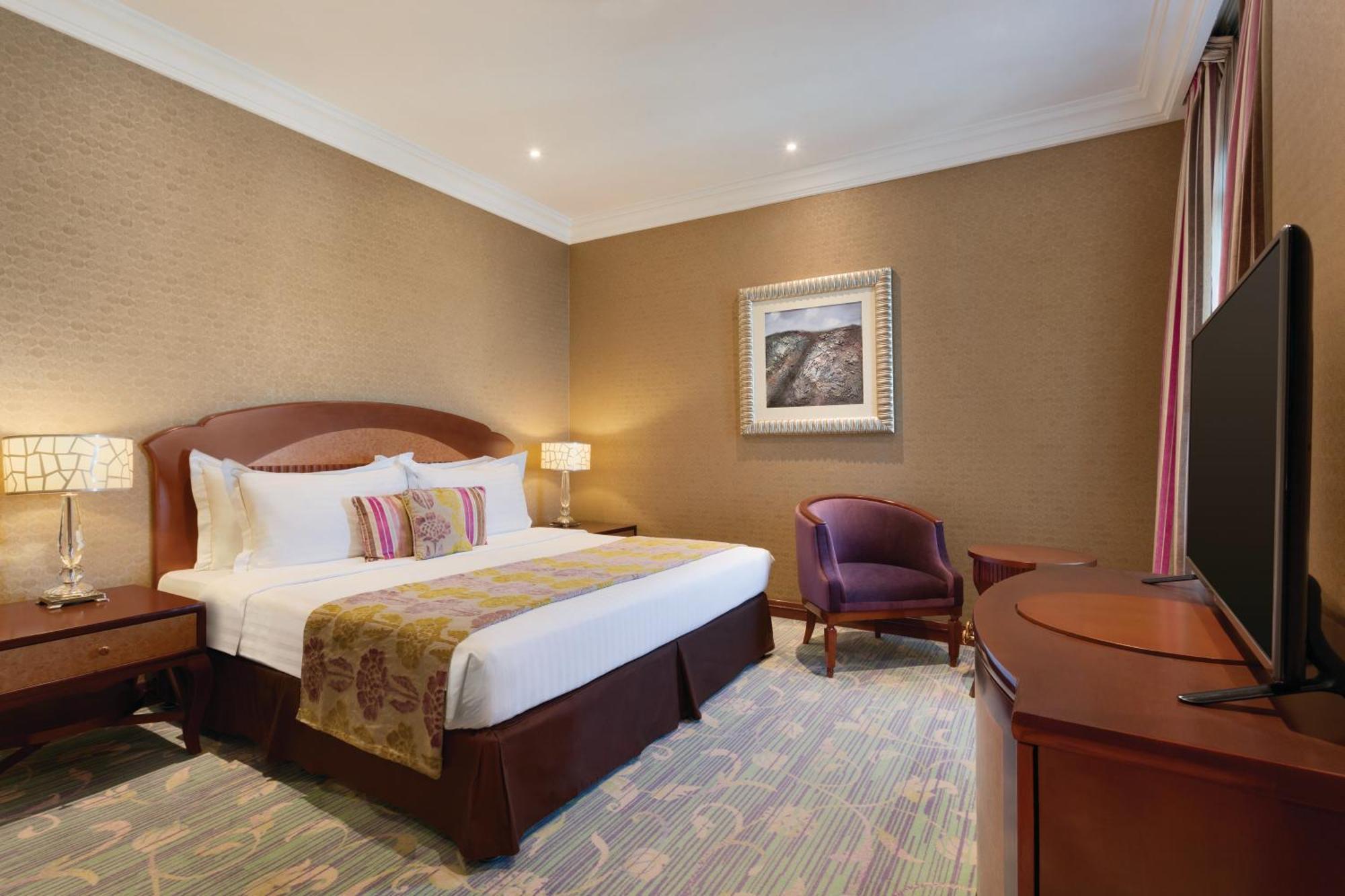 Grand Regency Doha, Trademark Collection By Wyndham Екстериор снимка The image shows a hotel room that features a cozy and elegant design. There's a large bed with white sheets and colorful decorative pillows. Next to the bed, there are nightstands with lamps. A comfortable armchair is positioned in the corner, and a 