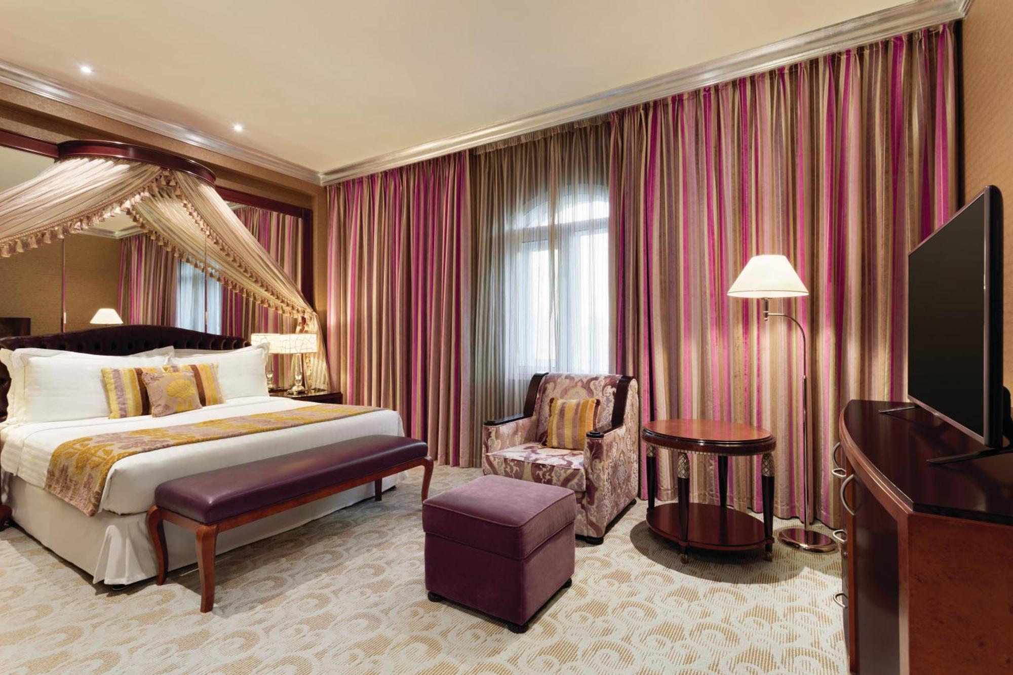 Grand Regency Doha, Trademark Collection By Wyndham Екстериор снимка The photo shows a stylish hotel room. It features a large bed with decorative pillows and a draped canopy above it. The room has elegant striped curtains in shades of pink and purple. There is a small seating area with a plush chair and ottoman, alon