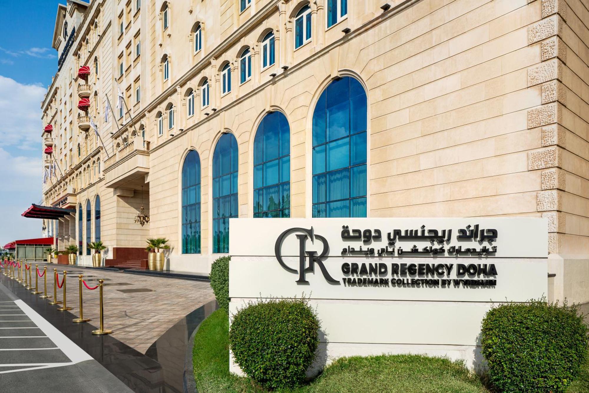 Grand Regency Doha, Trademark Collection By Wyndham Екстериор снимка The photo shows the entrance of the Grand Regency Doha, a luxury hotel. The building features a distinctive architectural style with large arched windows. There is a prominent sign featuring the hotel's name in both English and Arabic. The surroundin