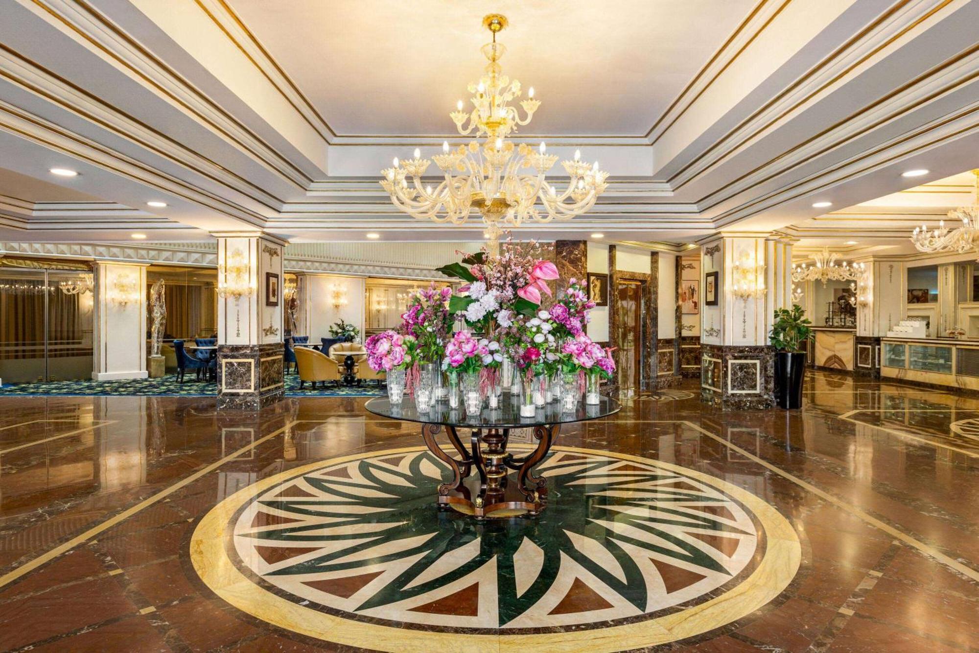 Grand Regency Doha, Trademark Collection By Wyndham Екстериор снимка The photo shows a luxurious hotel lobby featuring an elegant chandelier hanging from the ceiling. In the center, there is a large round table adorned with an arrangement of vibrant flowers. The floor showcases intricate marble patterns, and the overa