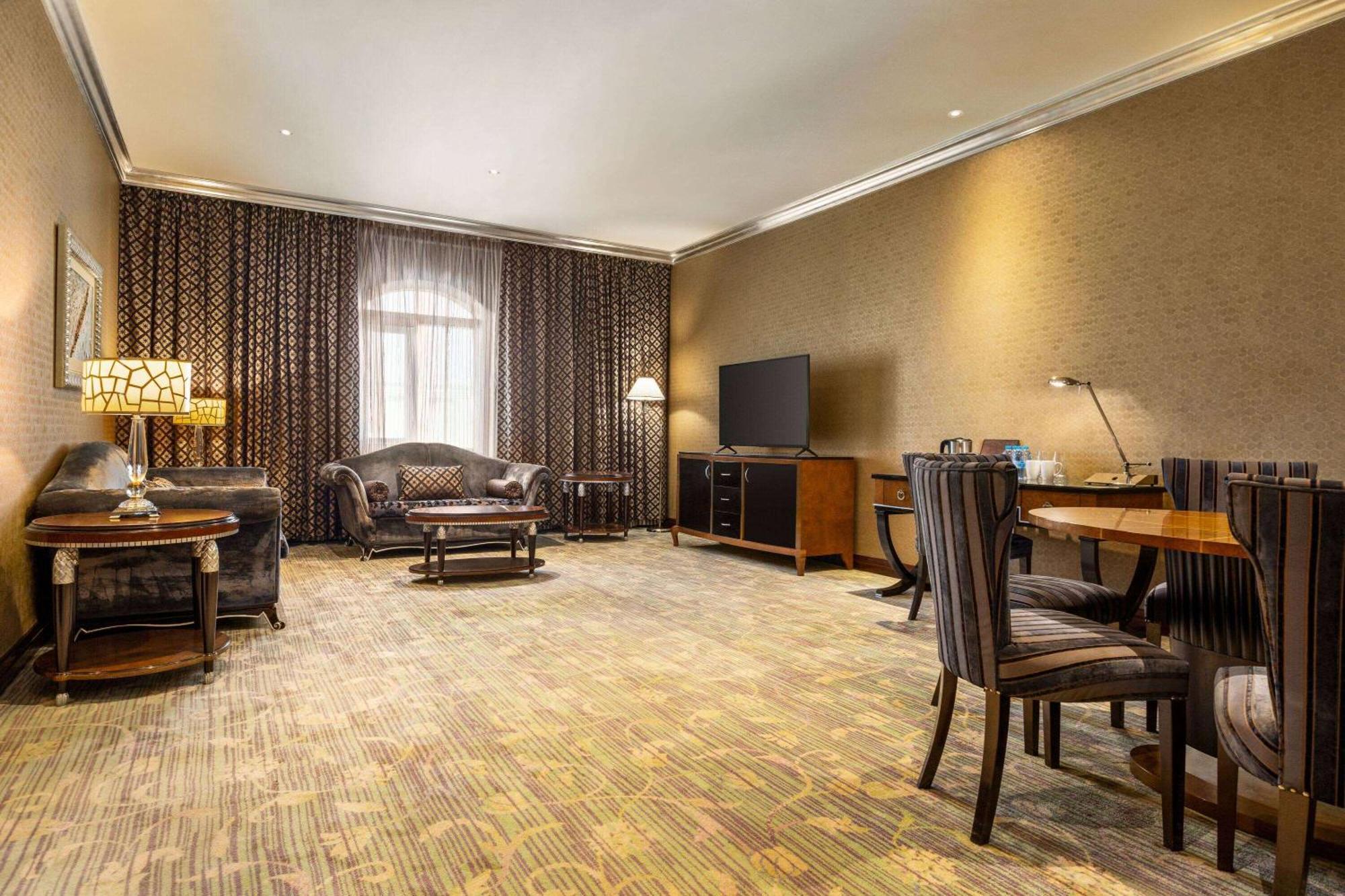 Grand Regency Doha, Trademark Collection By Wyndham Екстериор снимка The photo shows a spacious lounge or living area. It features a comfortable seating arrangement with a dark-colored sofa and armchairs, arranged around a coffee table. There's a dining table with matching chairs, suggesting it can accommodate meals o