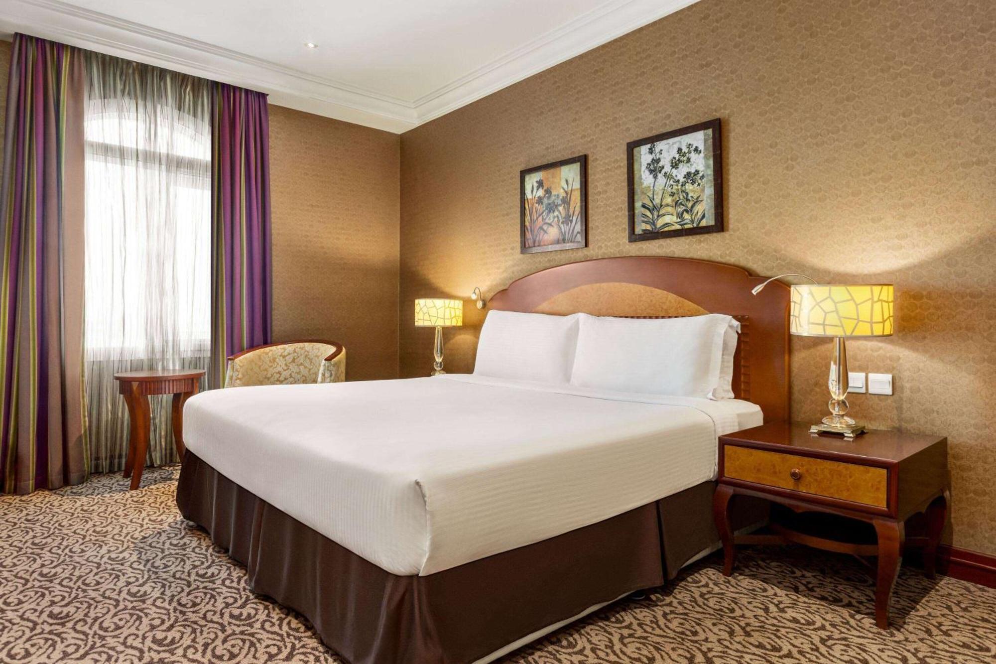 Grand Regency Doha, Trademark Collection By Wyndham Екстериор снимка The photo displays a comfortable hotel room featuring a large bed with white linens and a brown bed skirt. The room has a warm color palette, with textured brown walls and patterned carpet. There are two bedside lamps providing soft lighting next to 