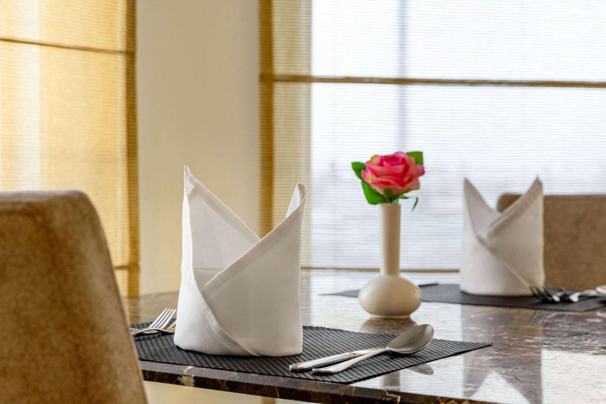 Grand Regency Doha, Trademark Collection By Wyndham Екстериор снимка The photo shows a neatly arranged dining table. On the table, there are two cloth napkins folded in a decorative way, one at each place setting. Silverware, including a knife and fork, is placed next to each napkin. In the center of the table, there 
