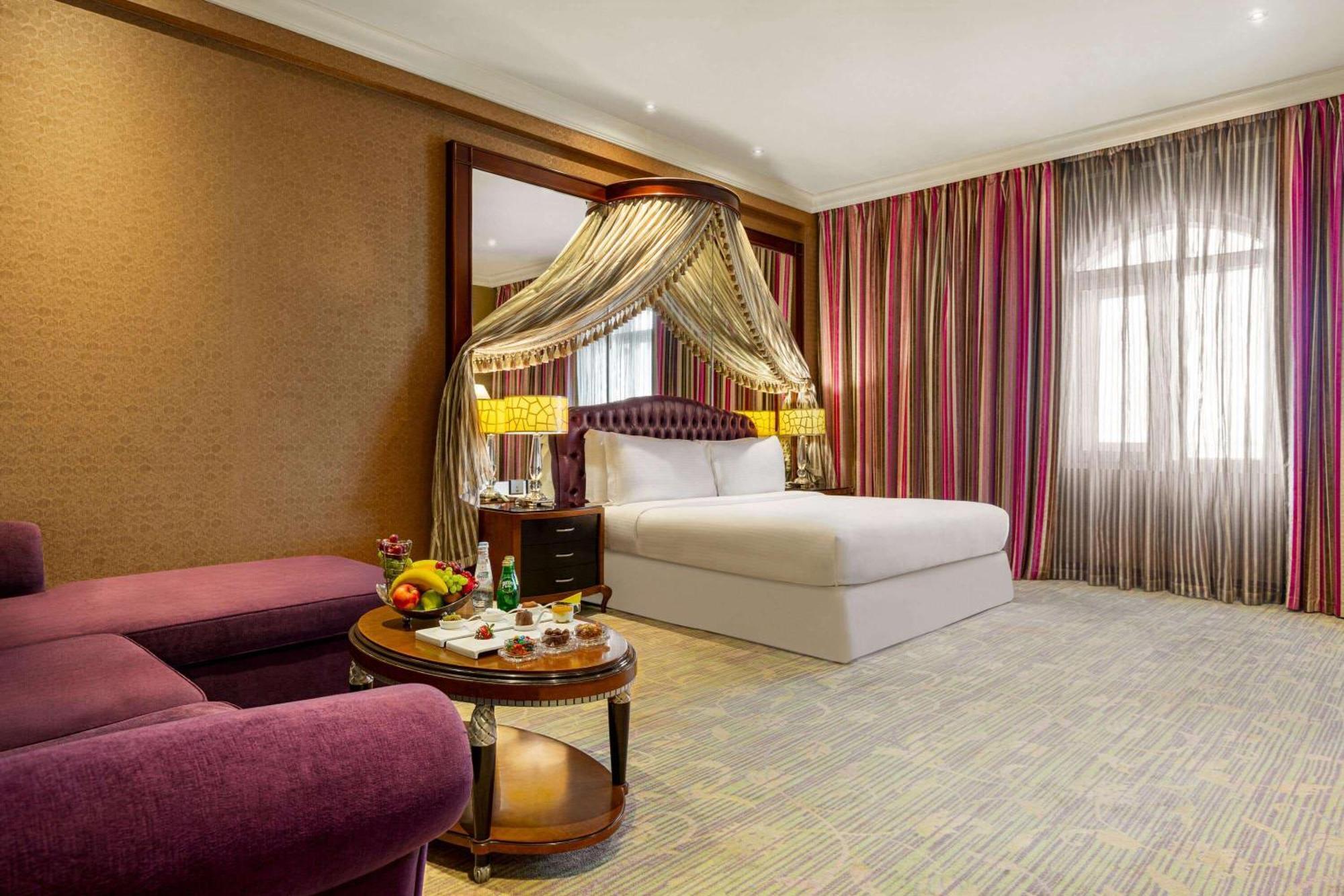 Grand Regency Doha, Trademark Collection By Wyndham Екстериор снимка The photo shows a luxurious hotel room featuring a king-sized bed with a decorative canopy. The room is elegantly designed with a mix of rich colors and textures. To the left, there's a plush purple seating area with a small round table that displays