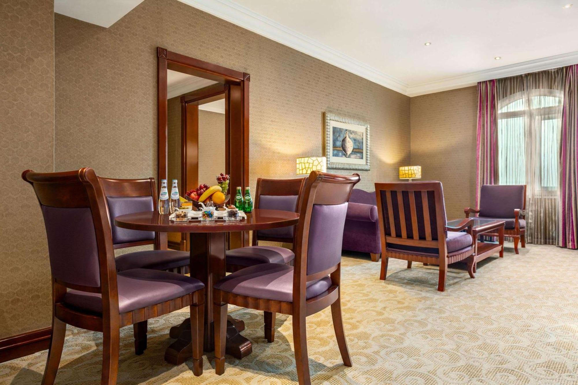 Grand Regency Doha, Trademark Collection By Wyndham Екстериор снимка The photo depicts a well-furnished hotel room or lounge area. In the foreground, there is a round table surrounded by four purple upholstered chairs, with a selection of fruits and beverages on the table. To the right, there are two wooden armchairs,